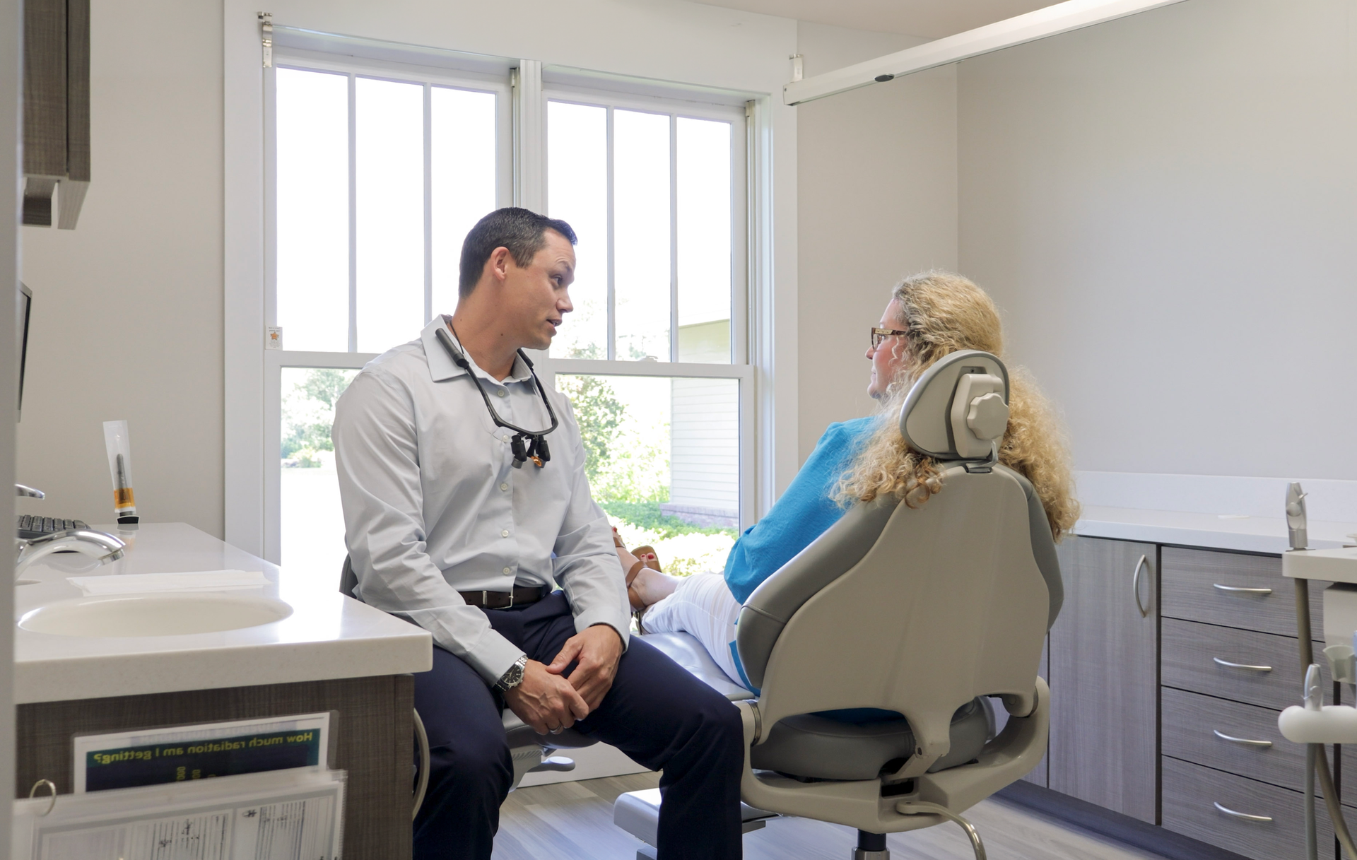 Woman sitting in a clinic waiting room, holding her cheek with a pained expression while a masked worker sits behind her.