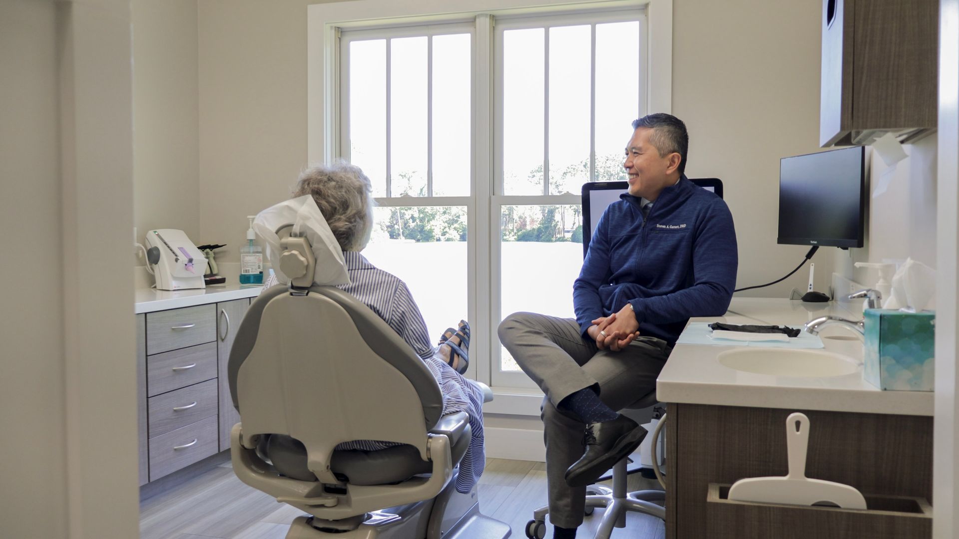 Smiling person reclining at a dental chair, holding a mirror, with a dentist blurred in the foreground