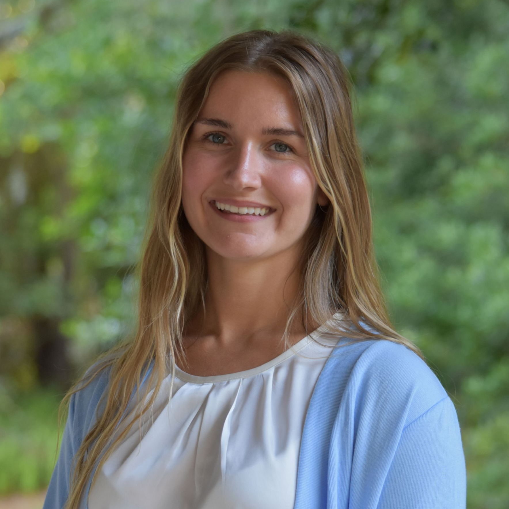 Smiling woman in a light blue outfit standing outdoors with a leafy green background