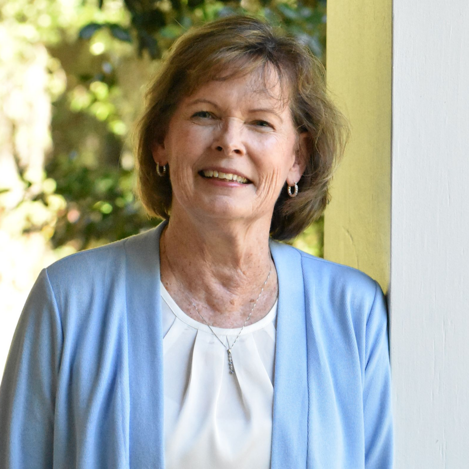 Woman standing by a yellow column outdoors, smiling in a light blue cardigan.