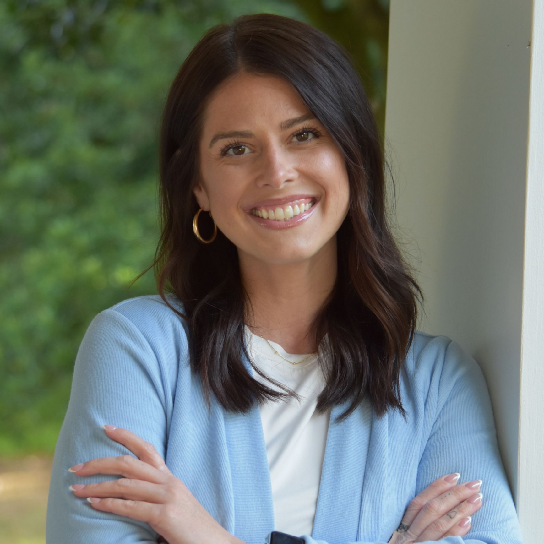 Woman smiling outdoors in a light blue cardigan, leaning against a porch column
