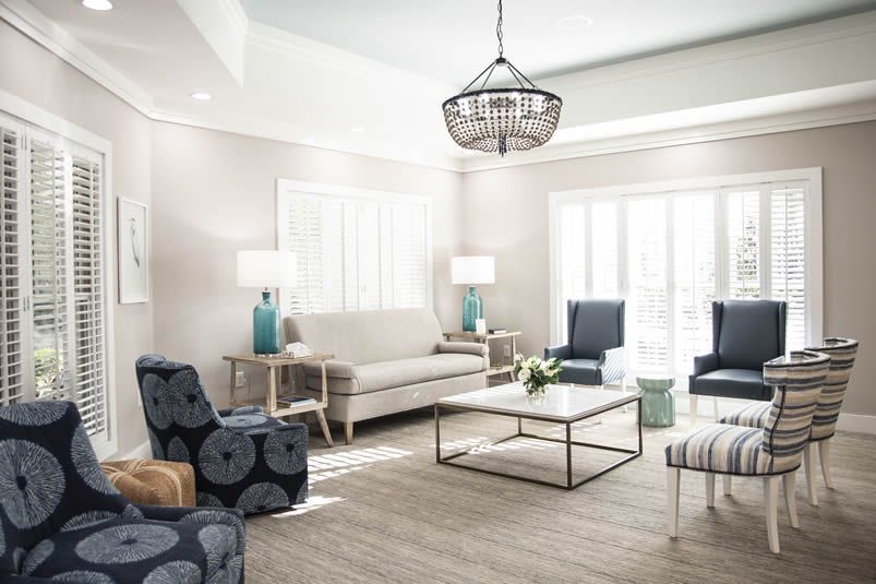 Bright living room with white shutters, blue patterned chairs, a beige sofa, and a glass coffee table