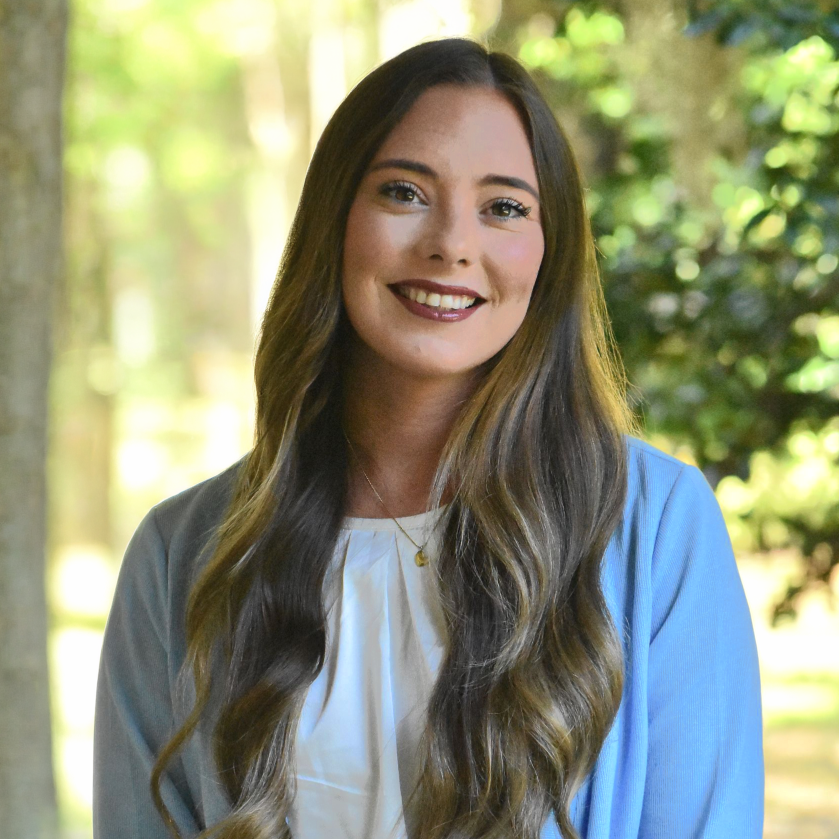 Smiling person with long wavy hair wearing a light blue cardigan outdoors in soft sunlight