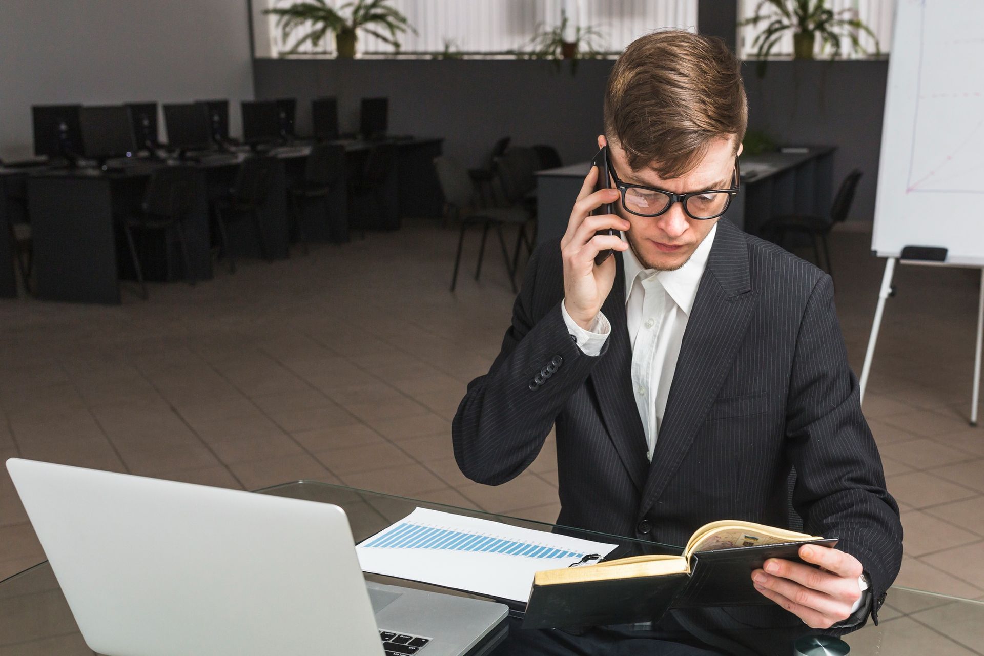 A man in a suit is sitting at a desk with a laptop and talking on a cell phone.