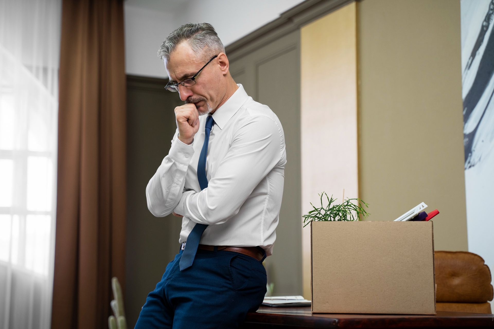 Man in office attire leans on desk, thoughtfully beside a box of belongings.
