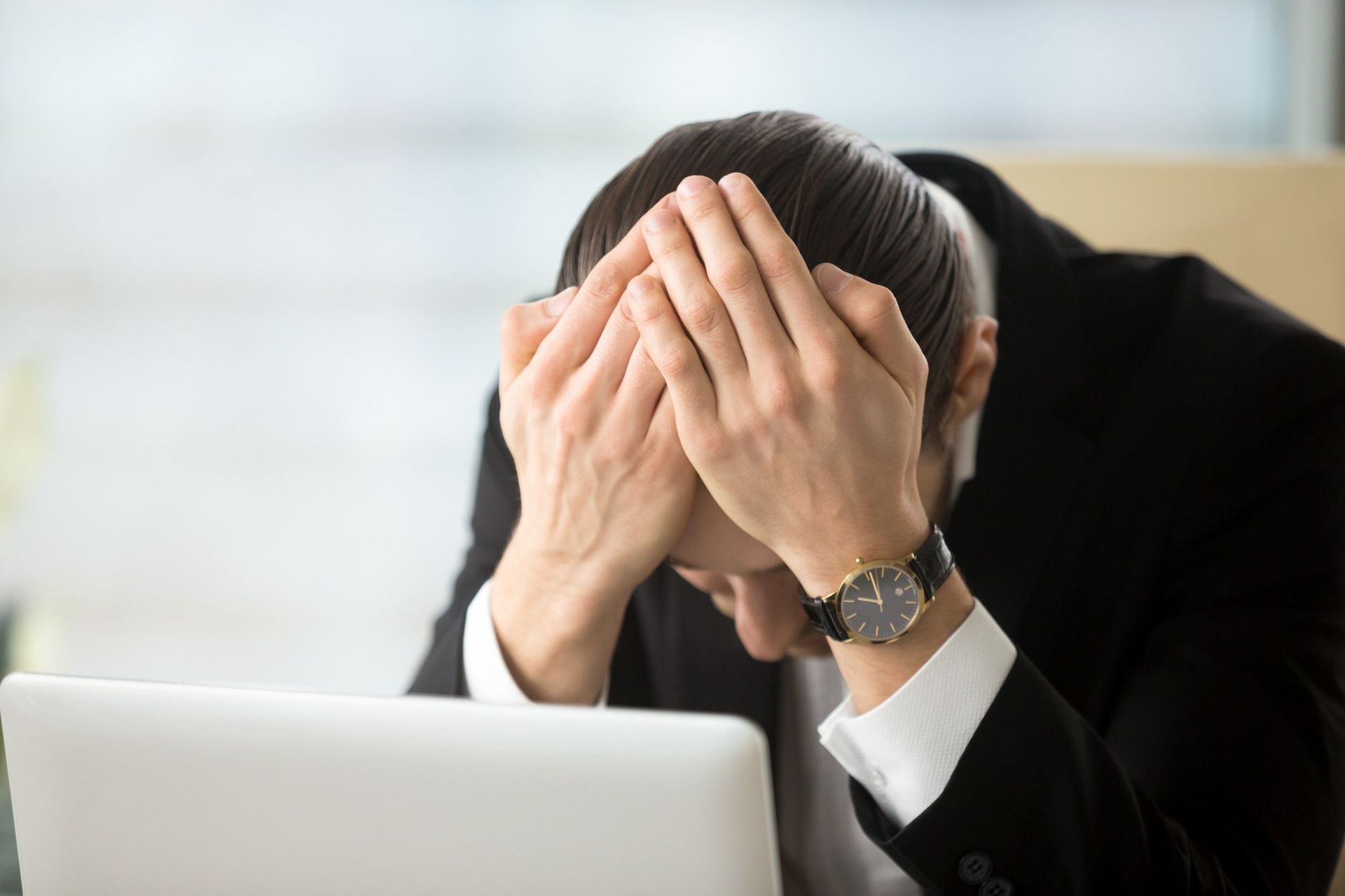 A man in a suit is covering his face with his hands while looking at a laptop.