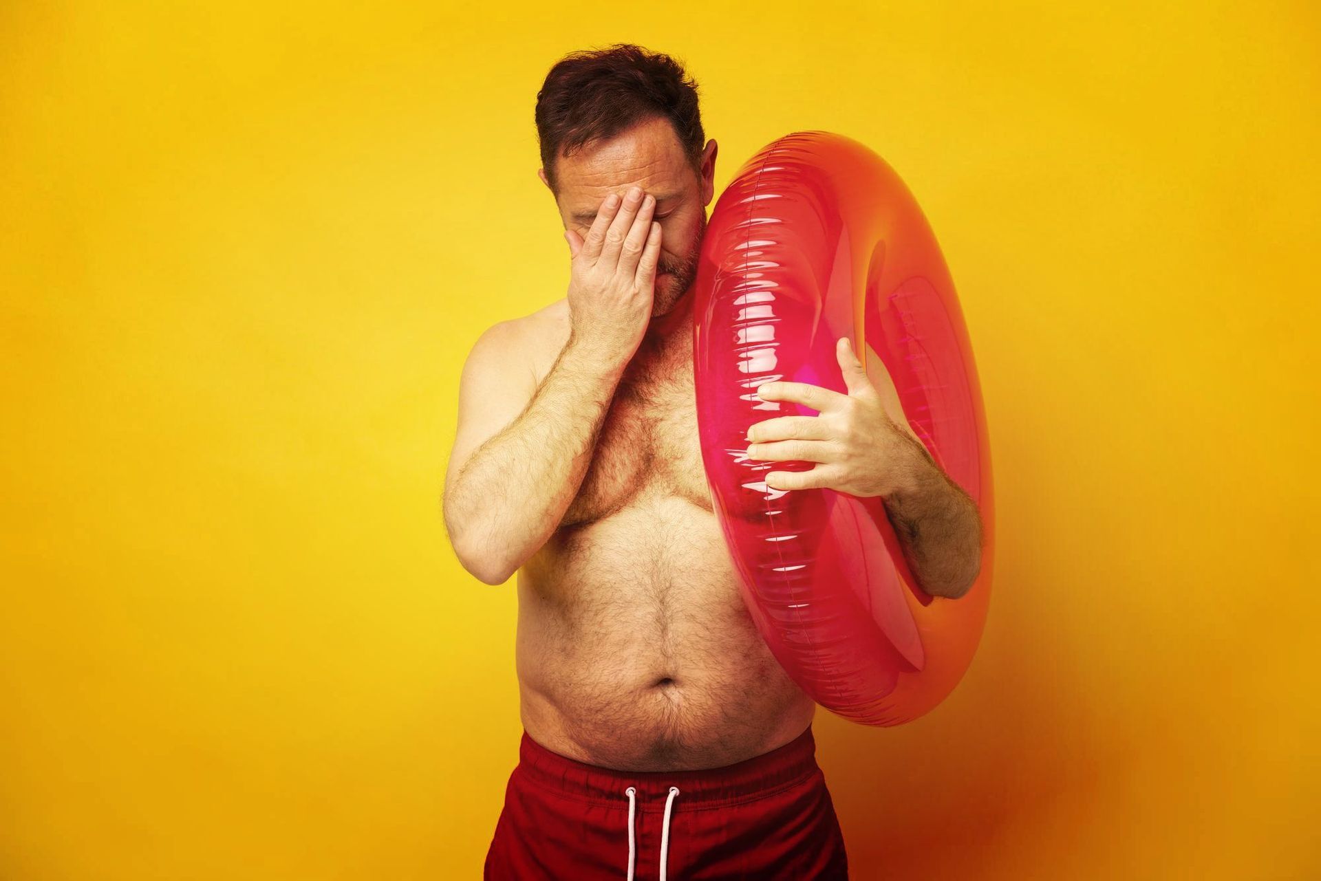 A shirtless man is holding an inflatable ring and covering his face.