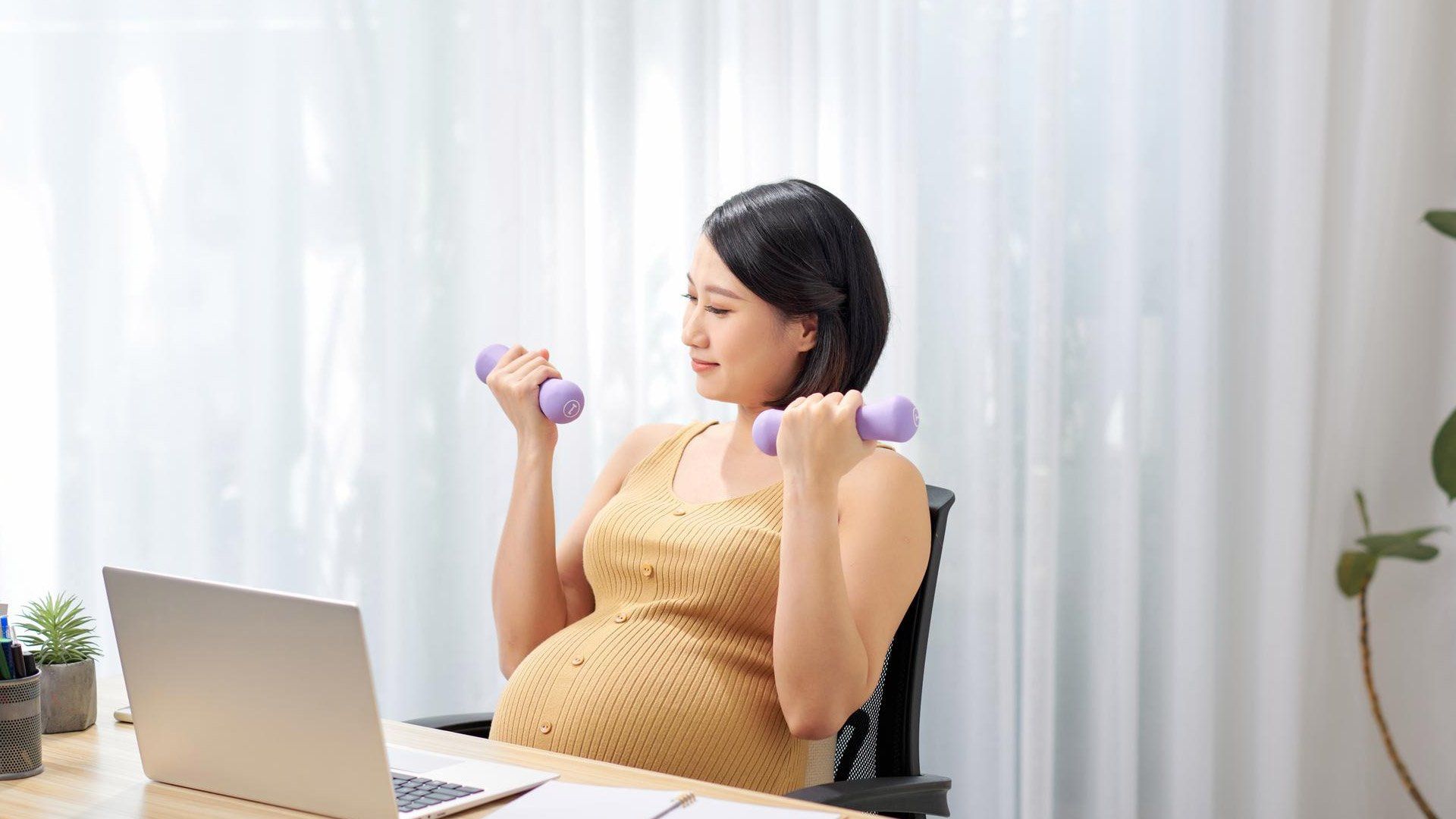 A pregnant woman is lifting dumbbells while sitting at a desk in front of a laptop.