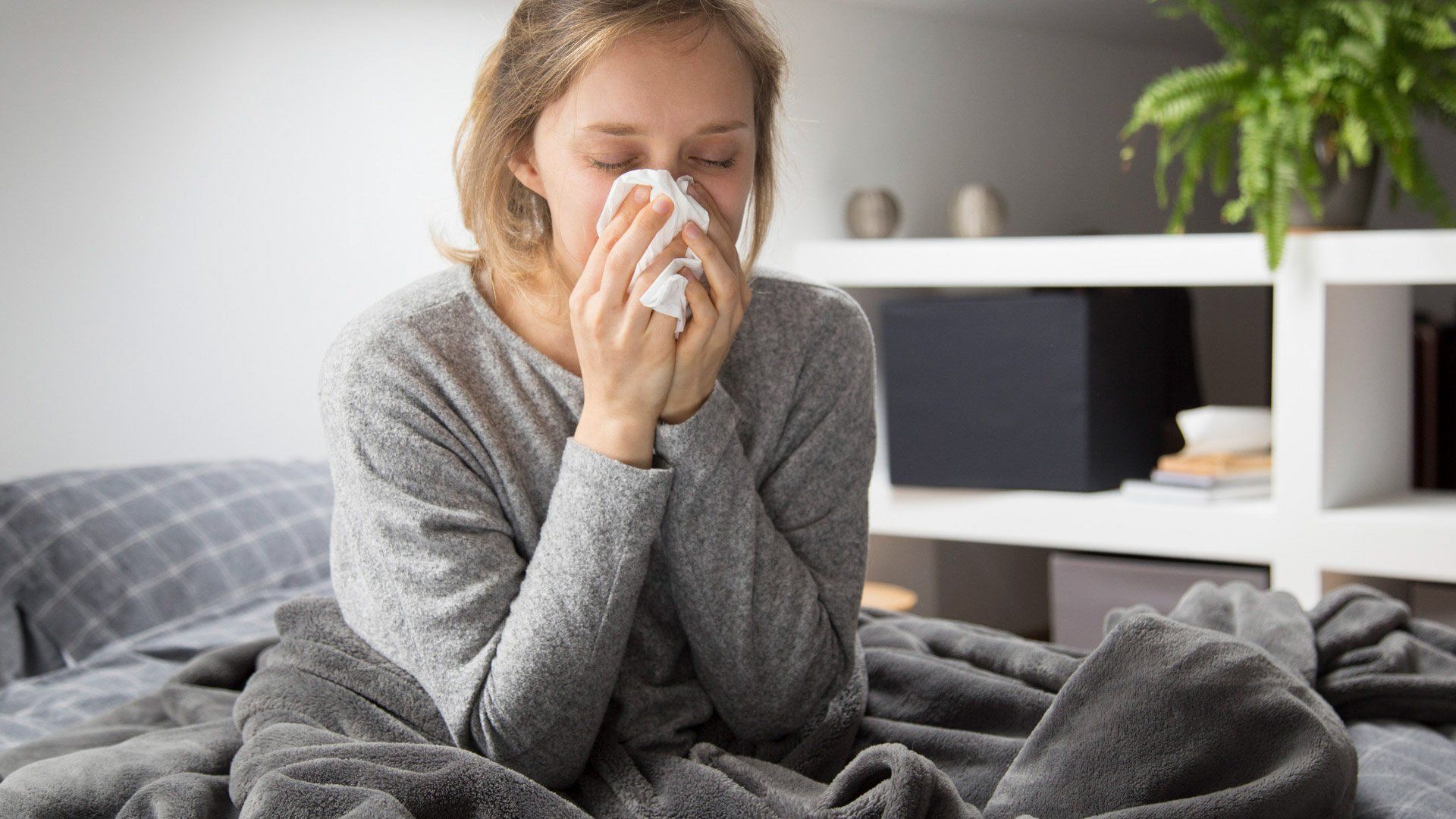 A woman is sitting on a bed blowing her nose into a napkin.