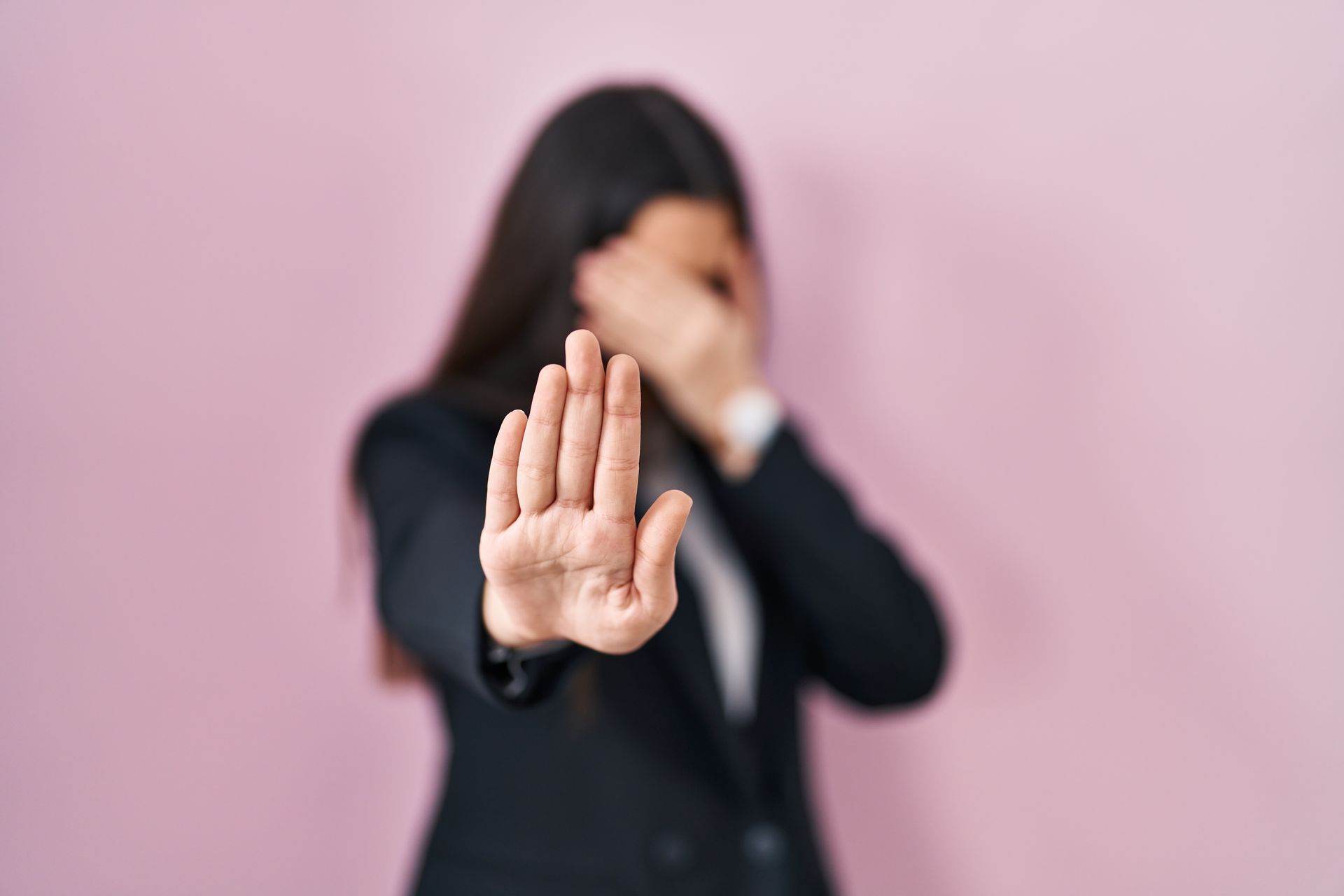 Woman in a dark blazer holds up a stop hand gesture against a pink background, face partially covered.