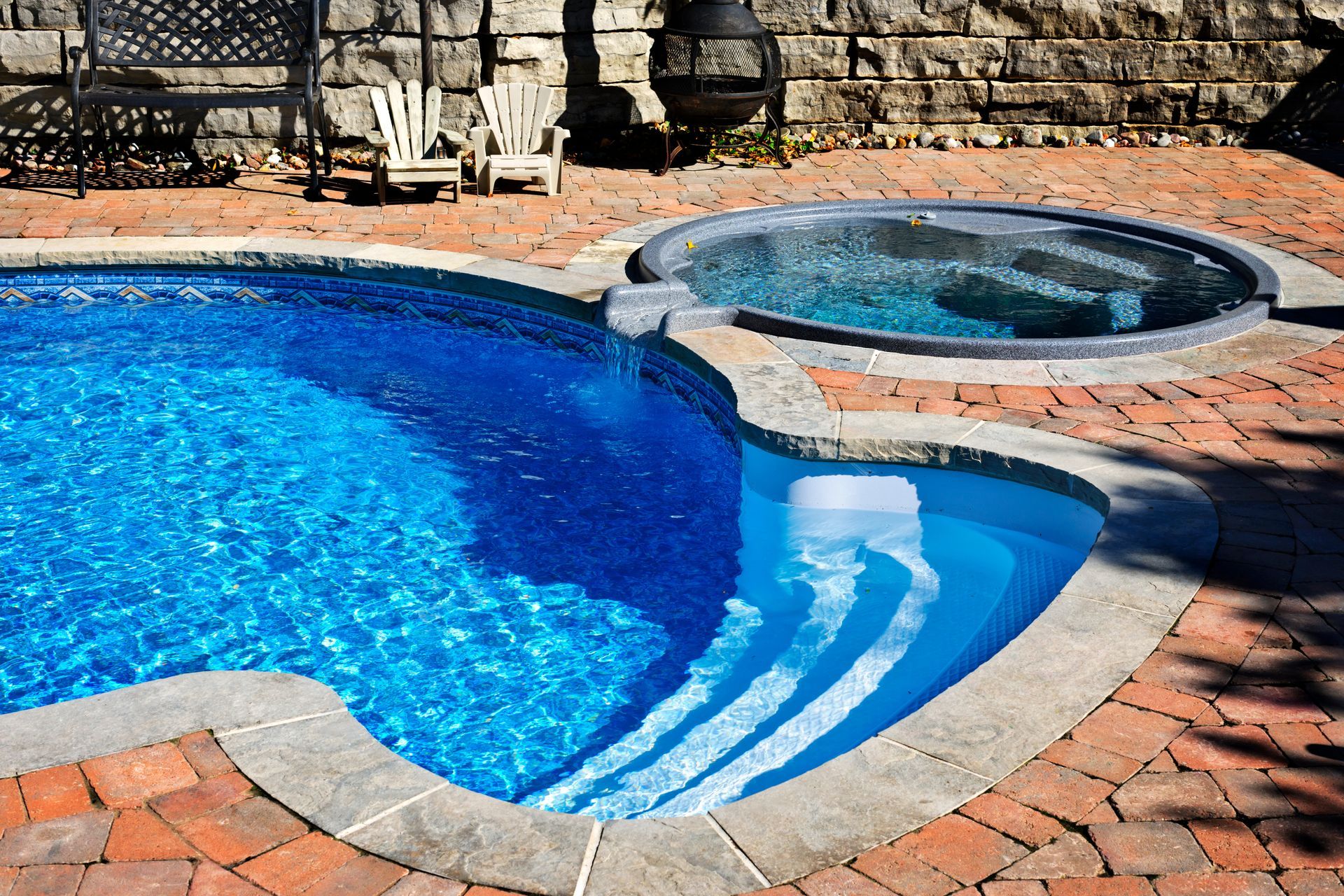 Backyard pool with hot tub spa, brick paver patio, stone wall, and Adirondack chairs.