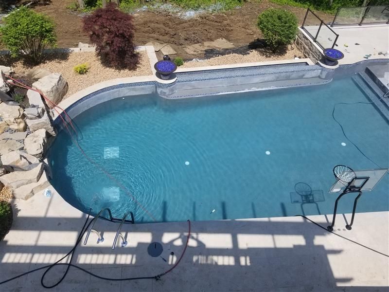 A pool with blue water and concrete deck. Landscaping includes rocks, plants, and trees.