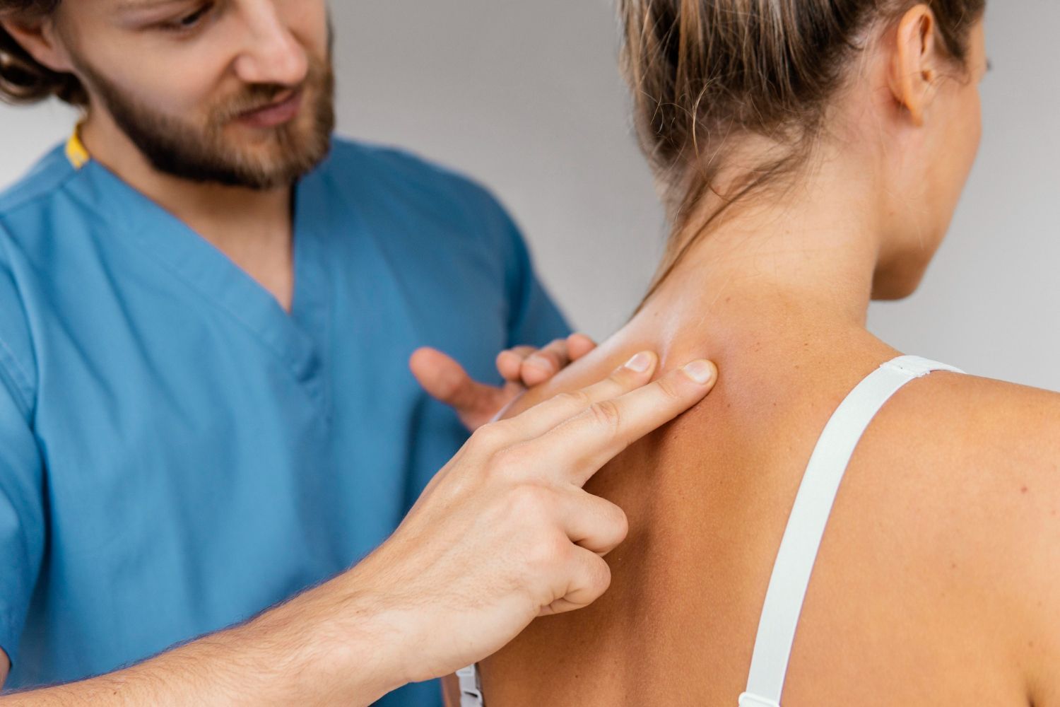 Physical therapist palpating a woman’s upper back and neck in a clinic, wearing blue scrubs