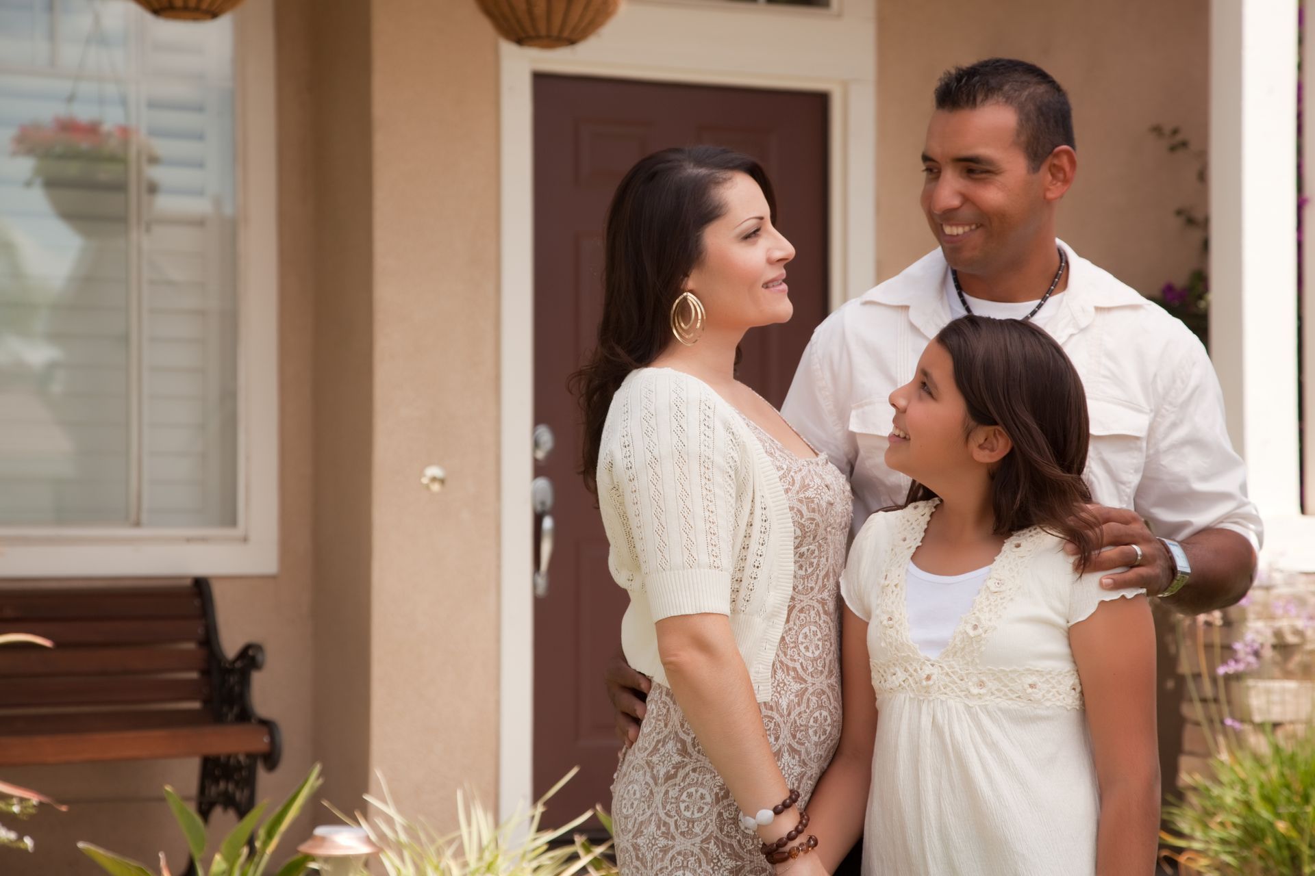 Family of three standing outside a house, smiling and holding hands, with the father’s arm around them.