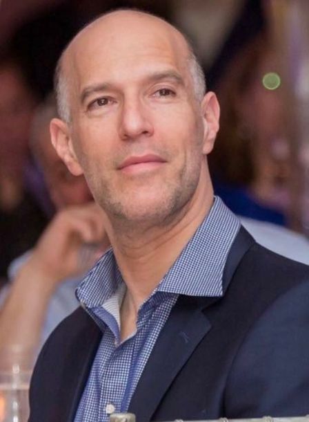Man in a navy blazer and patterned shirt posing at an event, with a blurred crowd behind him.