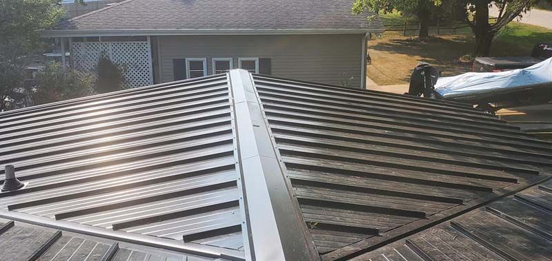 View of a metal roof with a central ridge, and a house in the background.
