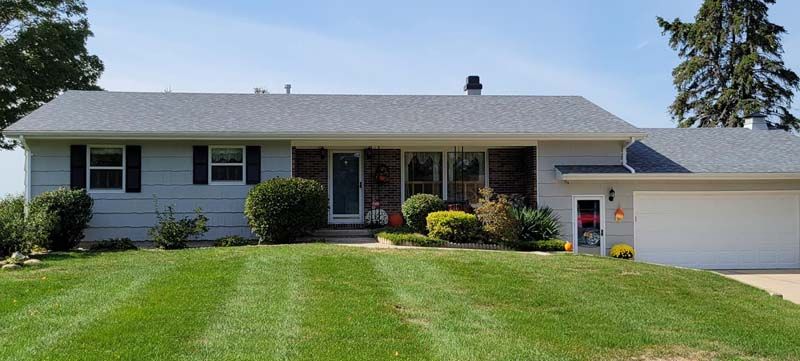 A light blue ranch-style house with black shutters, a gray roof, and a well-manicured lawn under a blue sky.