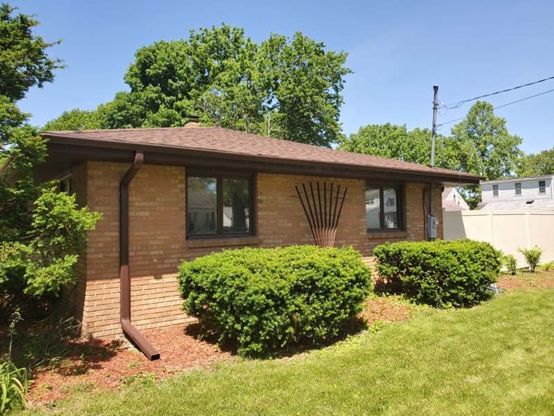 Brick house with brown roof and trim, green bushes, and lawn under a blue sky.