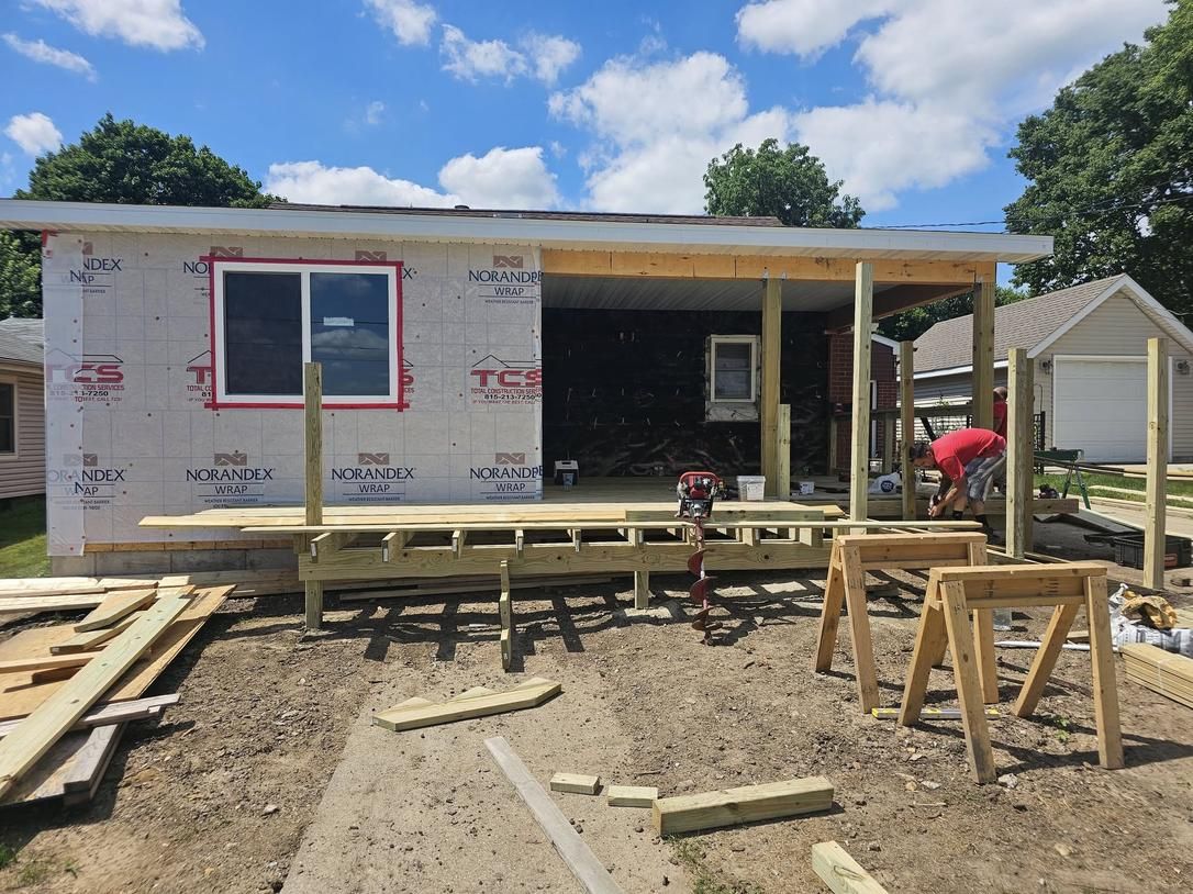 Construction of a small building with a deck. The walls are covered in sheathing, and a person works on the deck.
