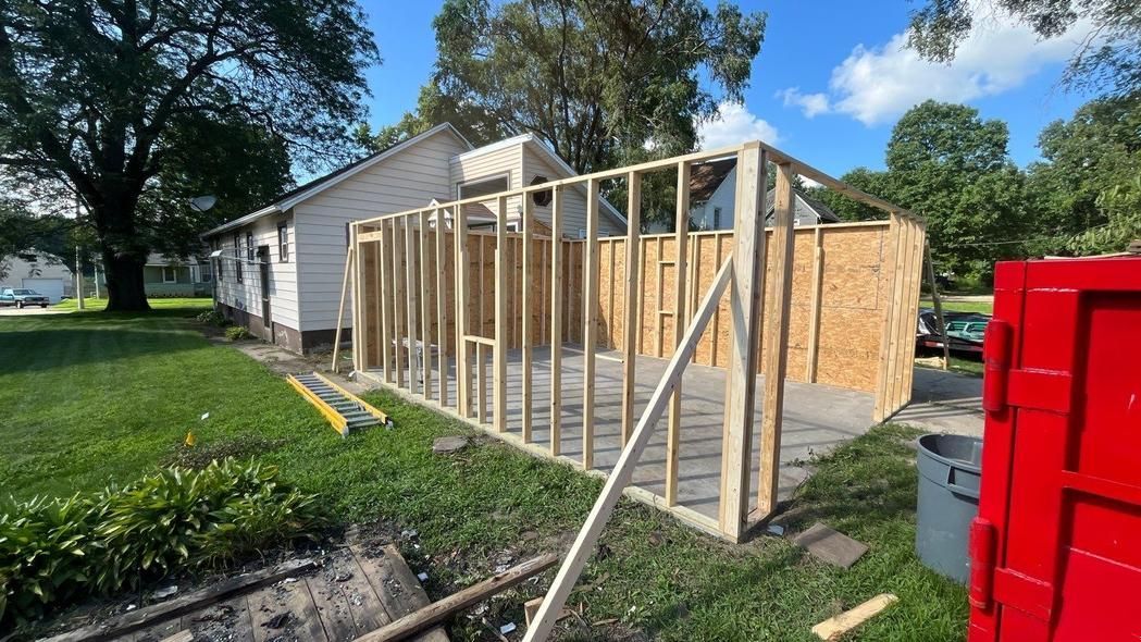 Construction framing for a home addition, with wood studs against the existing house and partial sheathing on a sunny day.