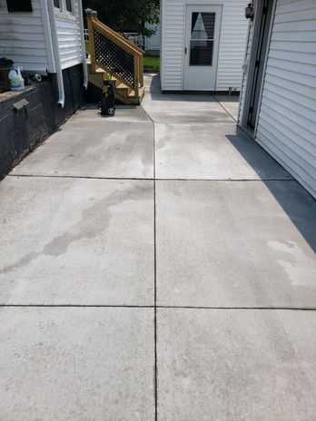 Concrete driveway with several gray squares, next to a white house and wooden stairs.