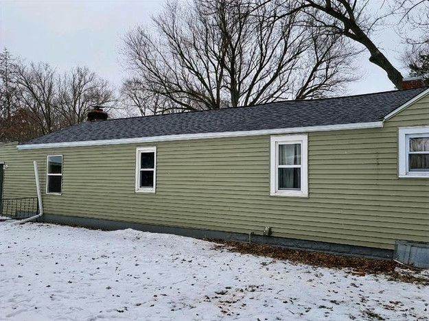 A single-story house with green siding, a dark gray roof, and white-framed windows, surrounded by snow.