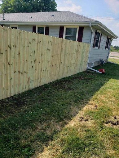Wooden fence bordering a house with red shutters. Green grass and blue sky.