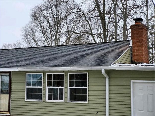 Green house with dark roof and brick chimney, three windows and a white door.
