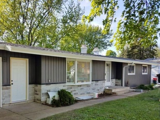 Gray and white ranch-style house with stone facade, white door, and green lawn in front of trees.