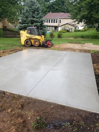 Newly poured concrete pad with a yellow skid steer in a yard setting.