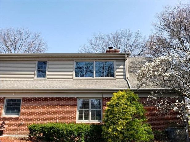 Two-story house with brick and beige siding. Windows reflect the sky. A blooming tree is on the right.