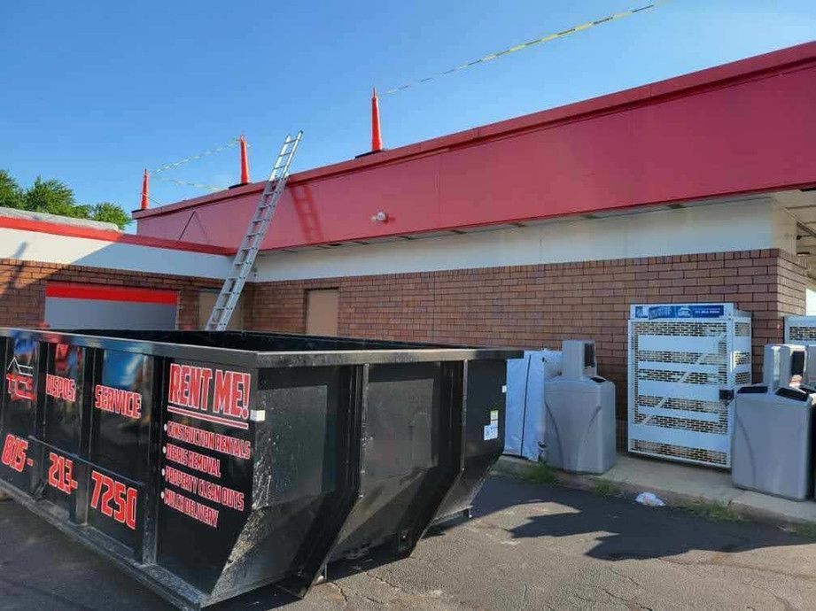 A black dumpster in front of a building with a red canopy; a ladder is propped against the roof.