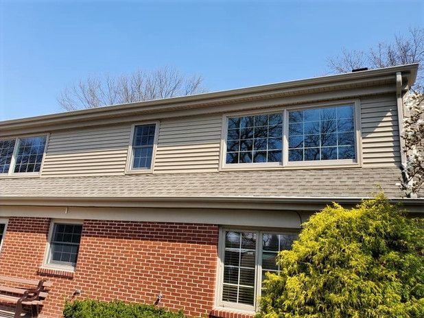 Two-story house with brick base, tan siding, multiple windows, and a clear blue sky.