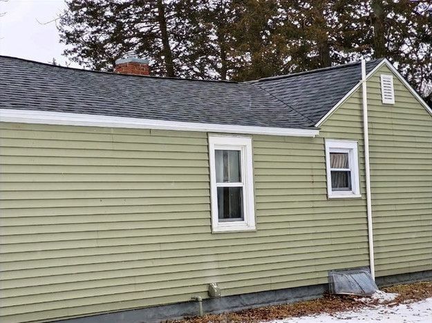 Green house with gray roof, white trim, two windows, and chimney. Snow on ground.