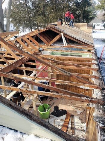 Roof partially demolished, exposing rafters. Workers in red jackets on top, snow on ground.