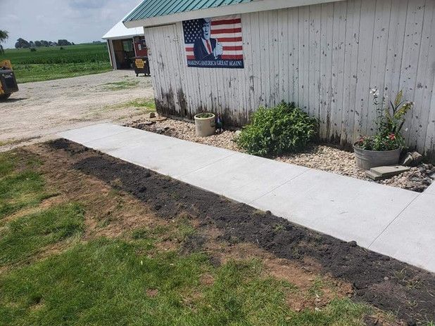 Concrete sidewalk next to a white building with an American flag, dirt patch in front of building, green grass.
