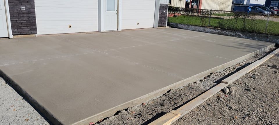 Newly poured concrete slab in front of a white building, with wooden form boards and surrounding dirt.