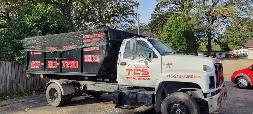 Dump truck with black bed and white cab, parked on a street. Company logo is visible. A red car is to the right.