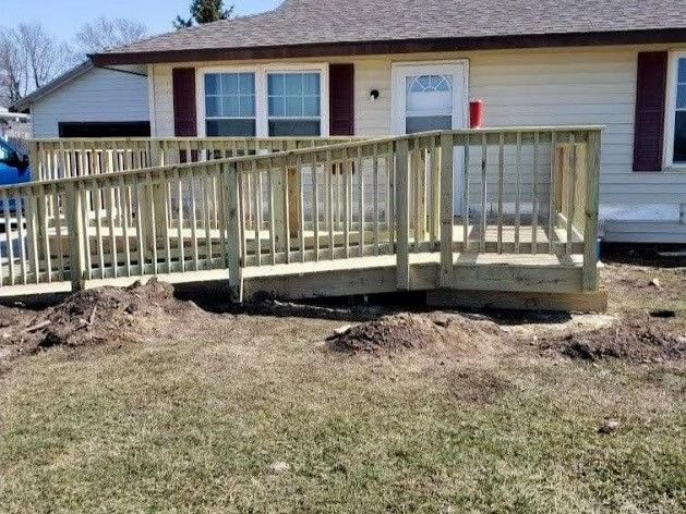 Wooden wheelchair ramp leading to the front door of a light-colored house. Brown roof and red shutters are visible.