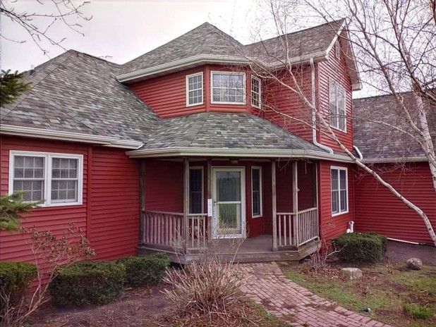Red house with porch and gray roof. Bushes and bare trees in front.