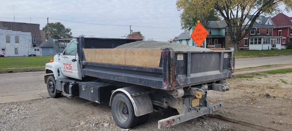 Dump truck filled with gravel on a road, orange construction sign in the background, houses along the street.