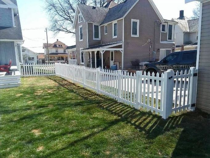 White picket fence surrounding a green grassy yard in front of a light brown house on a sunny day.