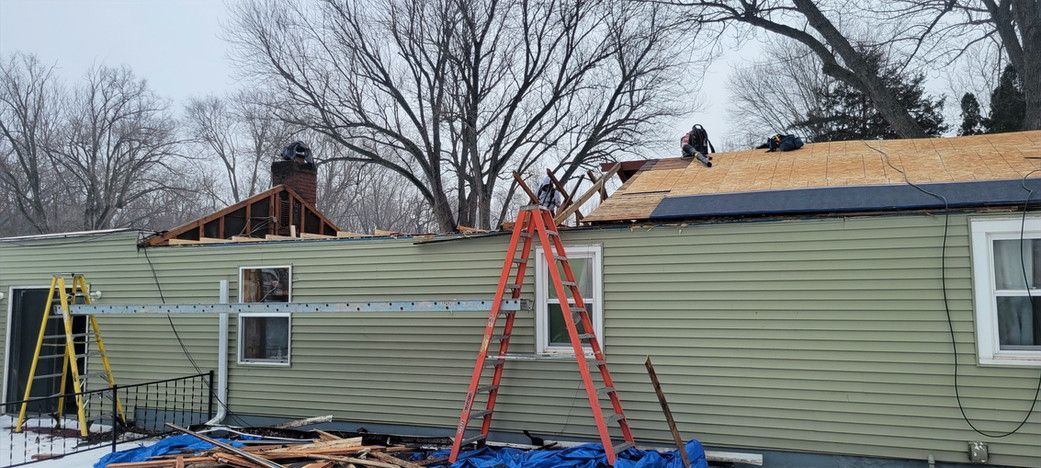 Roofing work in progress on a light green mobile home on a cold day.  Two workers on the roof, ladders and tarps visible.