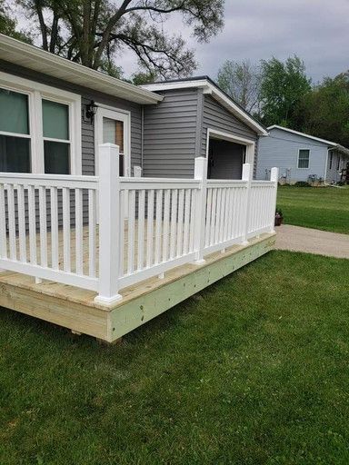 White railing encloses a wooden porch attached to a gray house, green grass in the foreground.