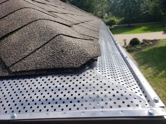Close-up of a roof with brown shingles and a perforated metal gutter guard, with a green yard in the background.