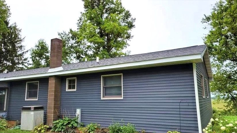 Dark blue house with a gray roof, brown chimney, and trees in the background under an overcast sky.