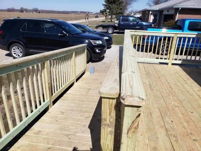 Wooden deck with ramp and railing, leading to a driveway with parked vehicles.