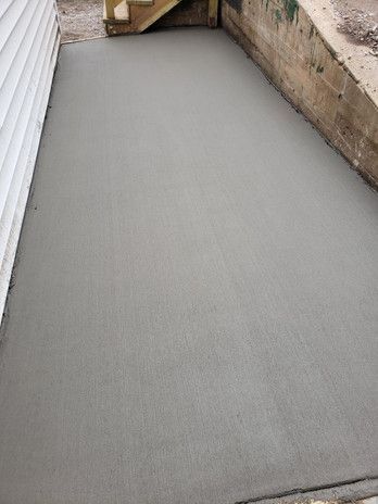Newly poured gray concrete patio next to white siding and a concrete wall. Wooden steps in the background.