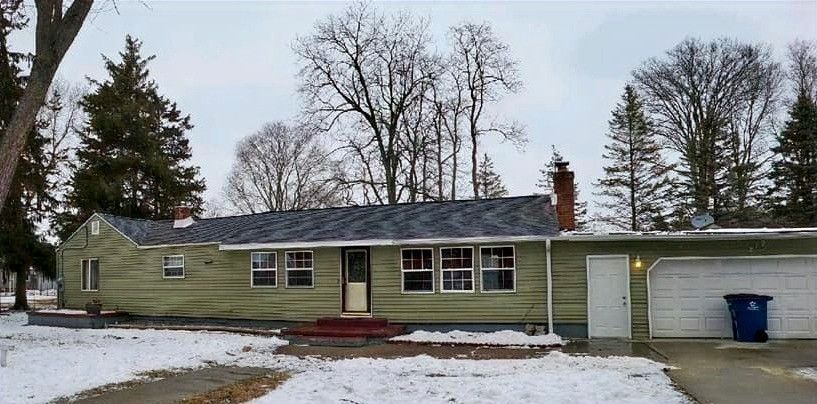 A single-story, green house with a gray roof, in a snowy setting with a garage and a blue trash can.