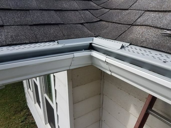 Damaged white gutter and roof shingles at a building's corner, with a ladder visible.